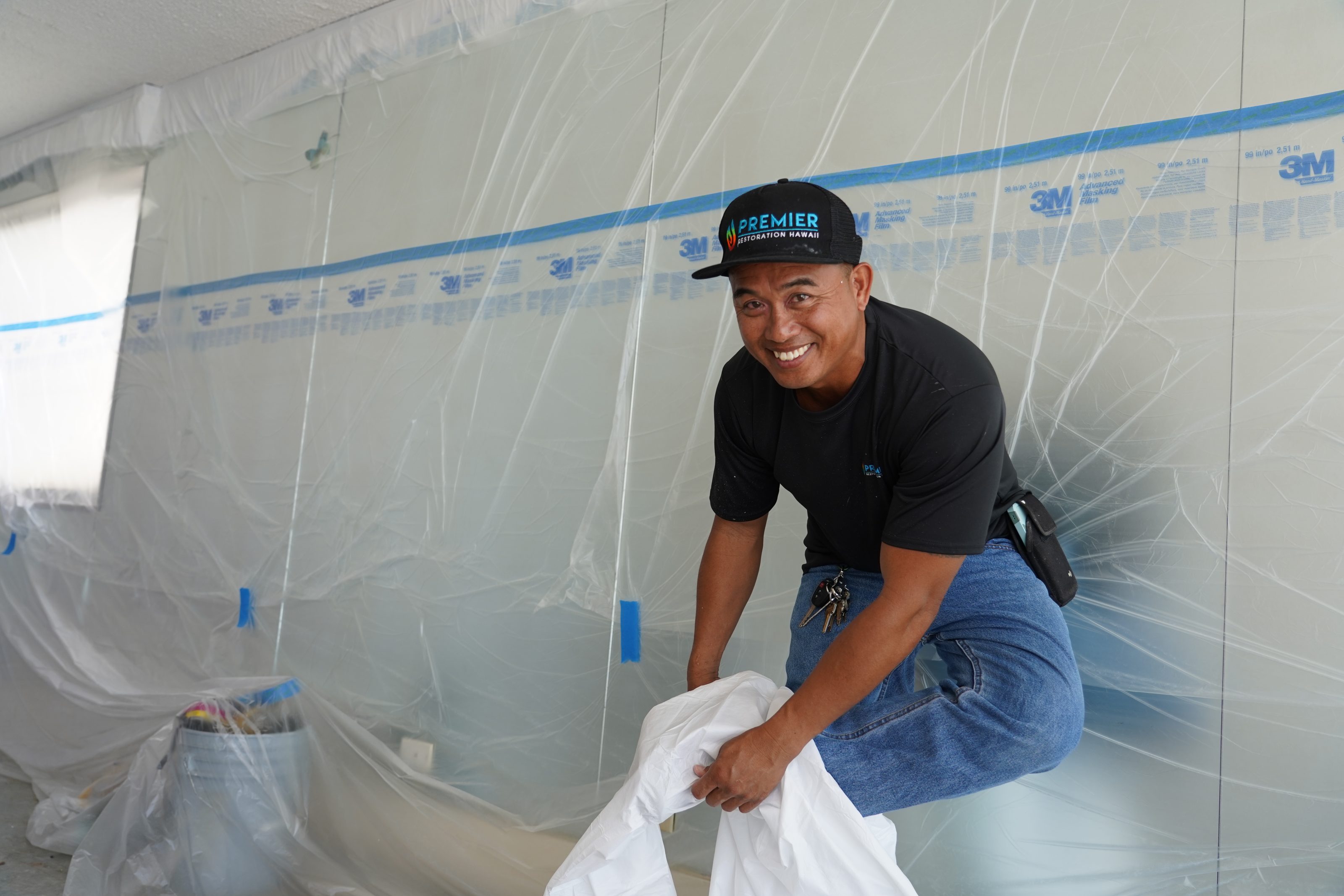 A man in a Premier Restoration Hawaii cap preparing a construction site with protective plastic sheeting.