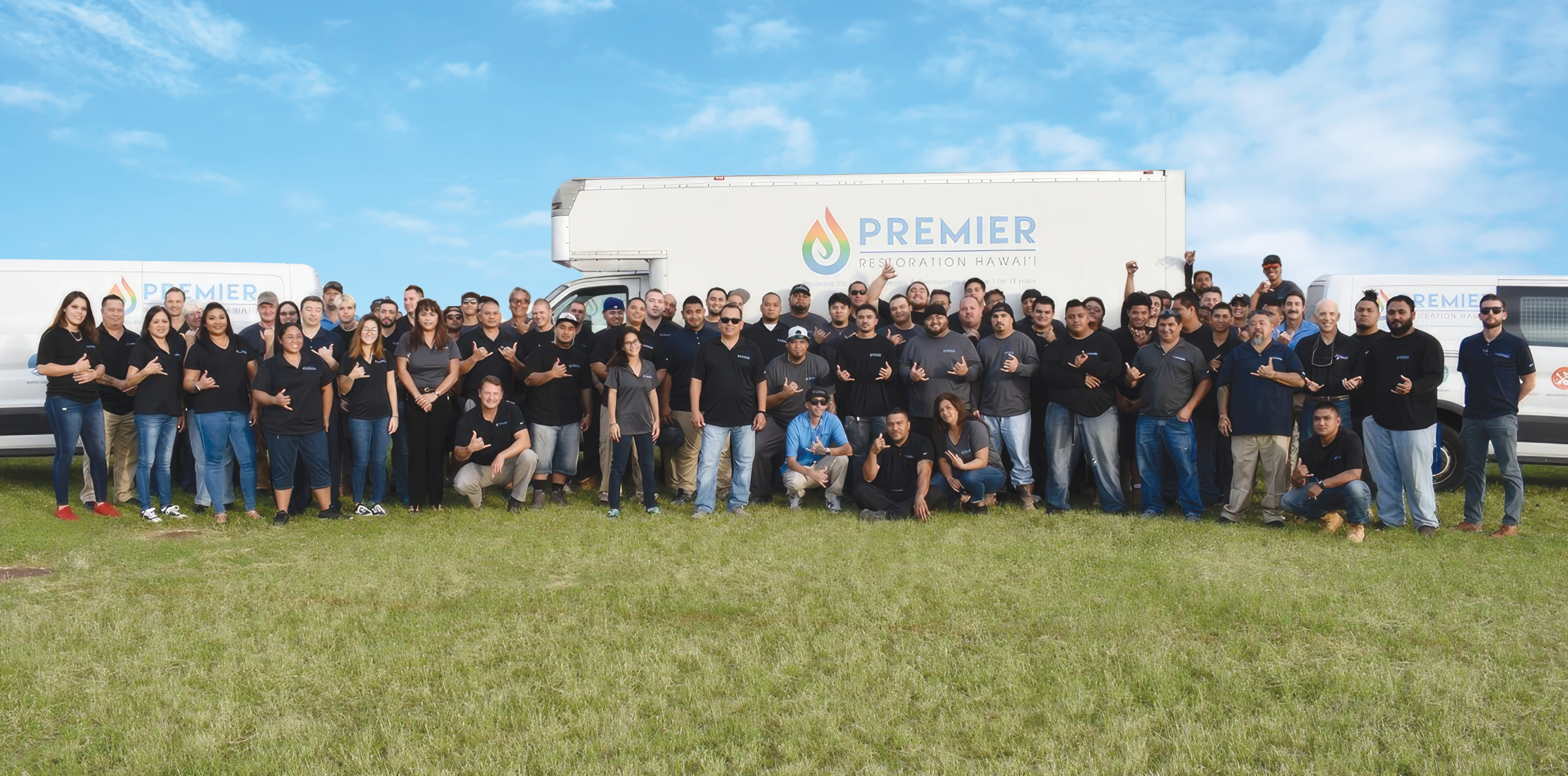 Team of Premier Restoration Hawaii gathers in front of company trucks on grassy field.
