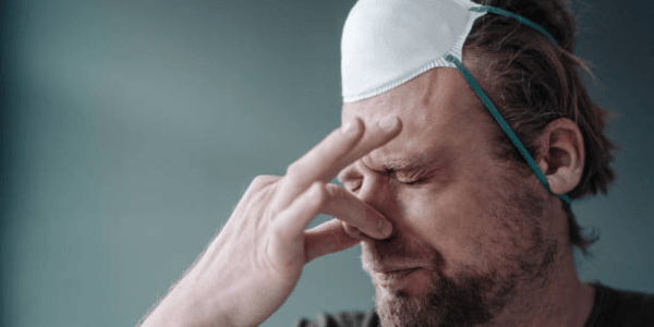 Man with mask on head pinching nose, appearing tired and stressed.