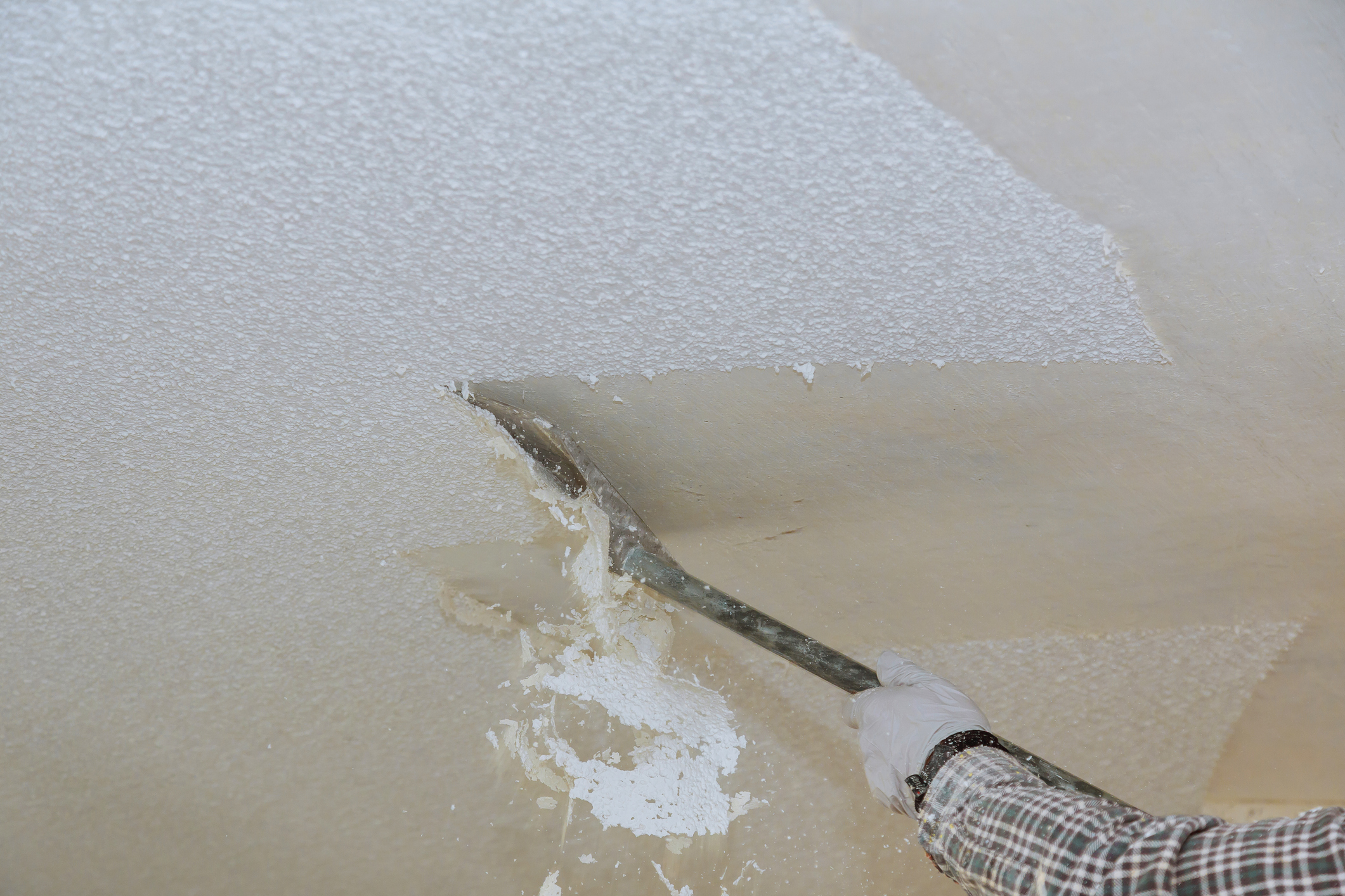 Person scraping popcorn ceiling off using a long-handled tool and wearing gloves.