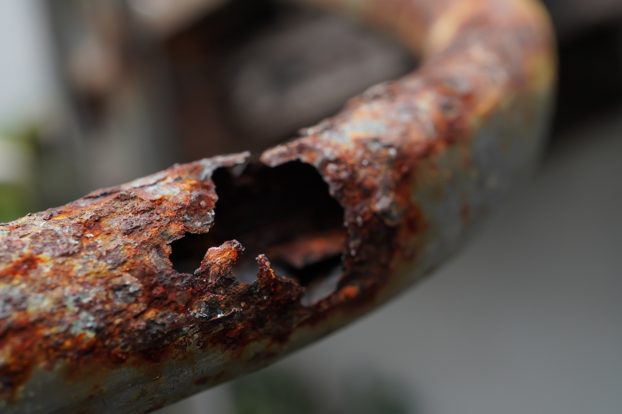 Close-up of a heavily corroded metal pipe with flaking orange rust and a jagged hole eaten through its wall.